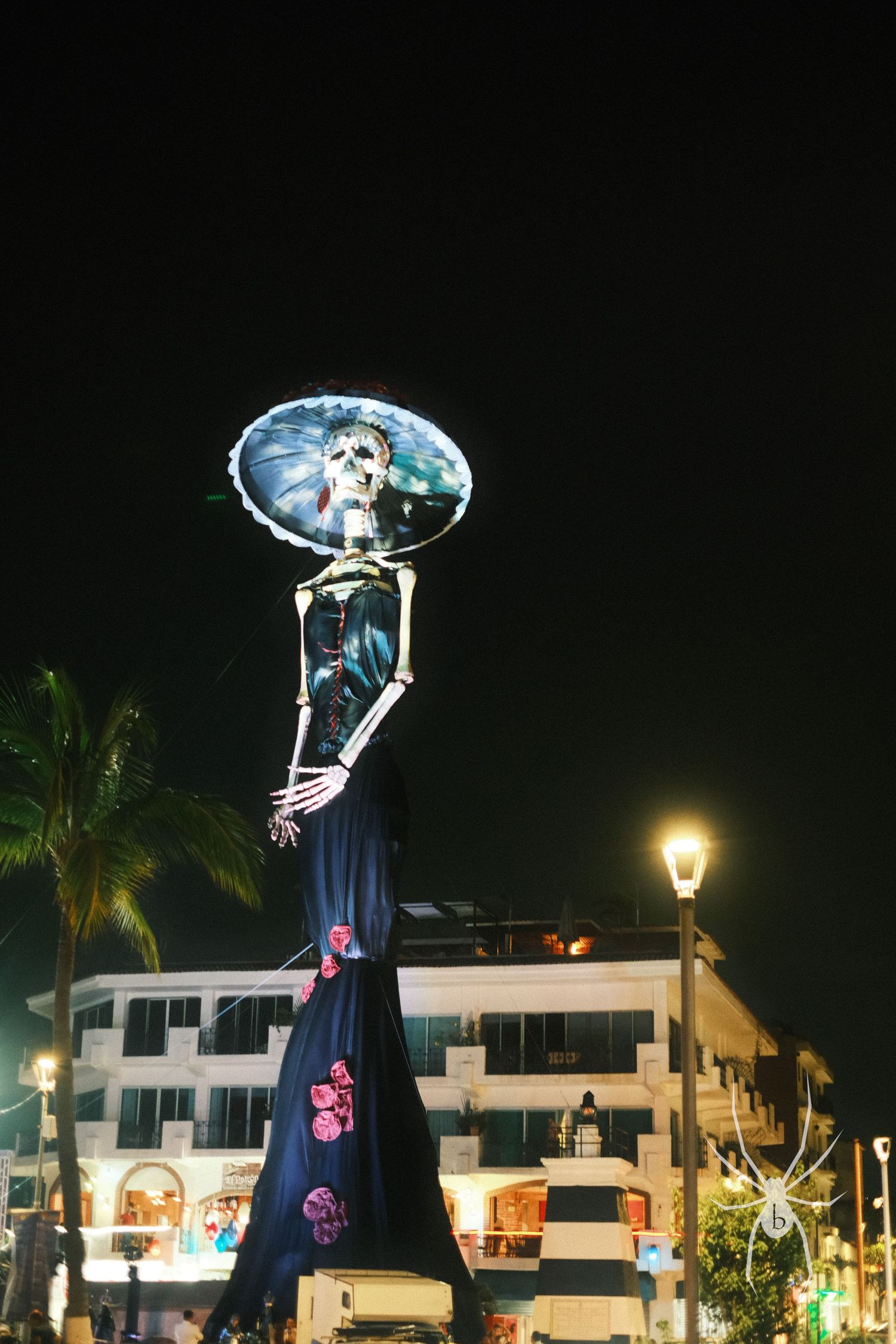 Tall skeleton decoration lit up at night with buildings and palm trees in the background.