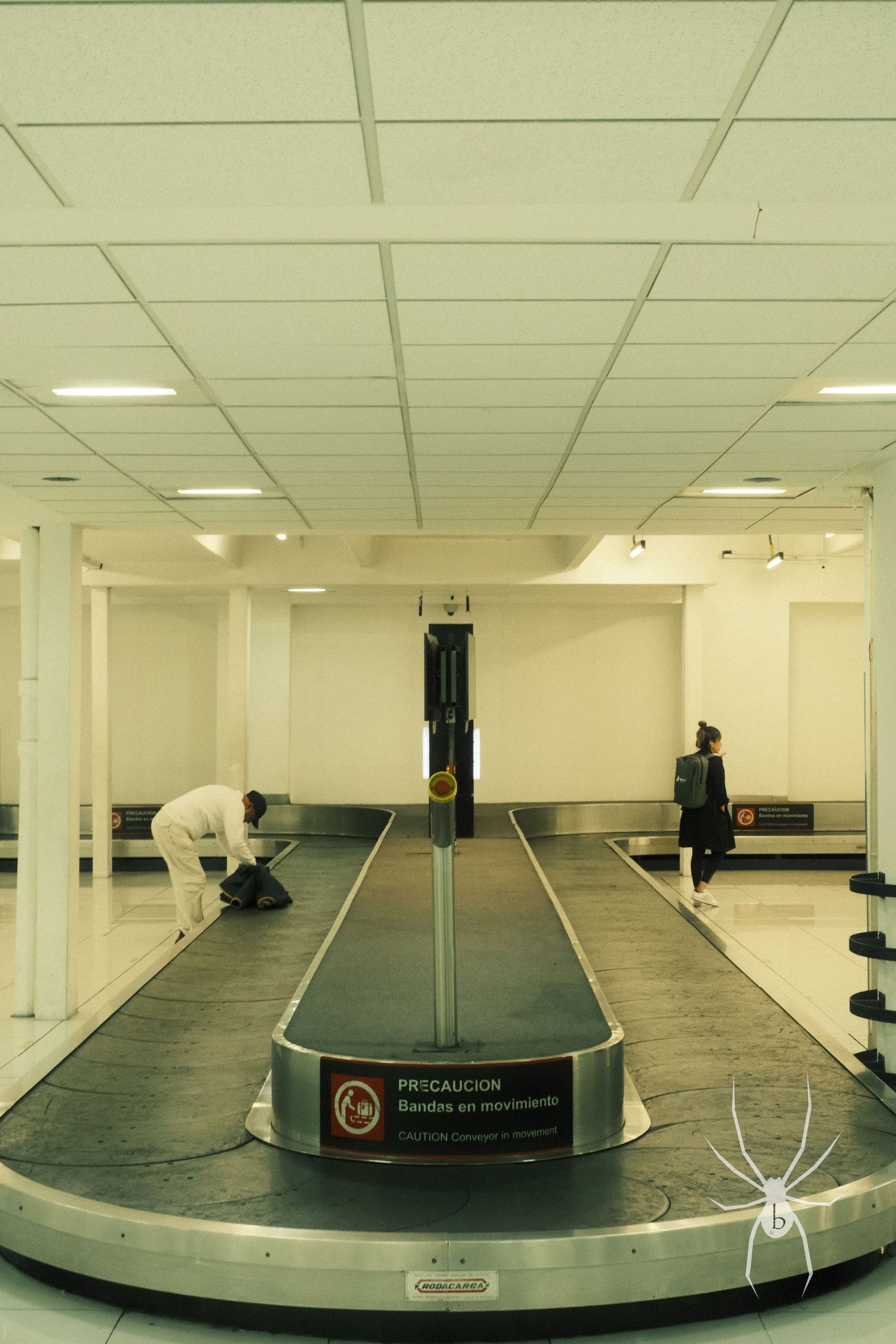 Empty baggage claim carousel in an airport terminal with white walls and fluorescent lighting.