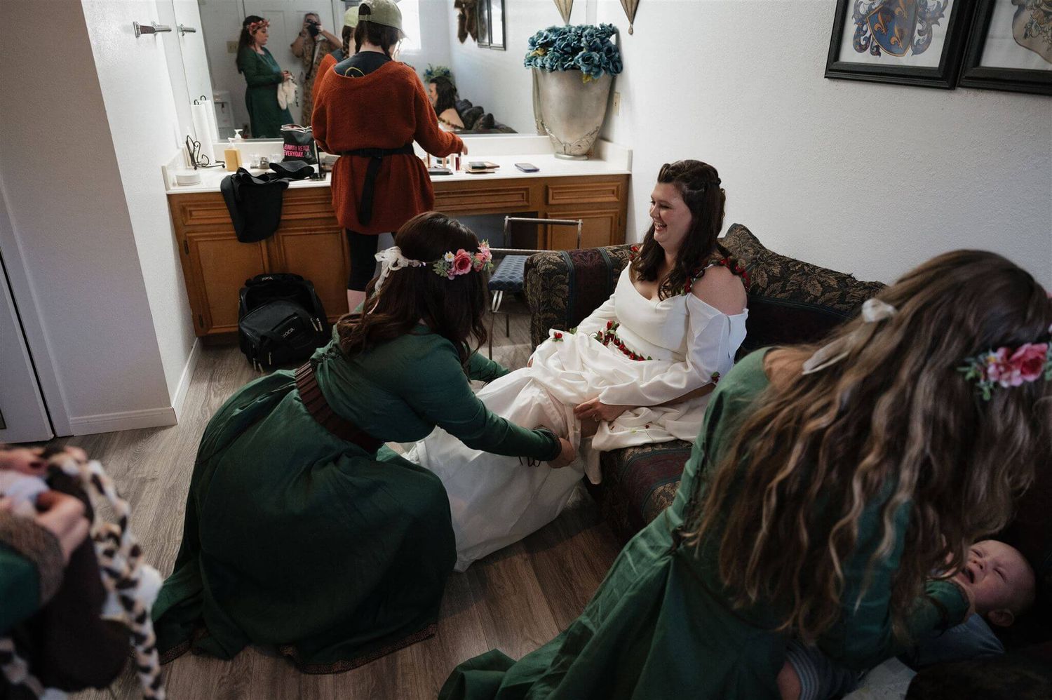 Several people in green period costumes gather around a chair in a dressing room setting.