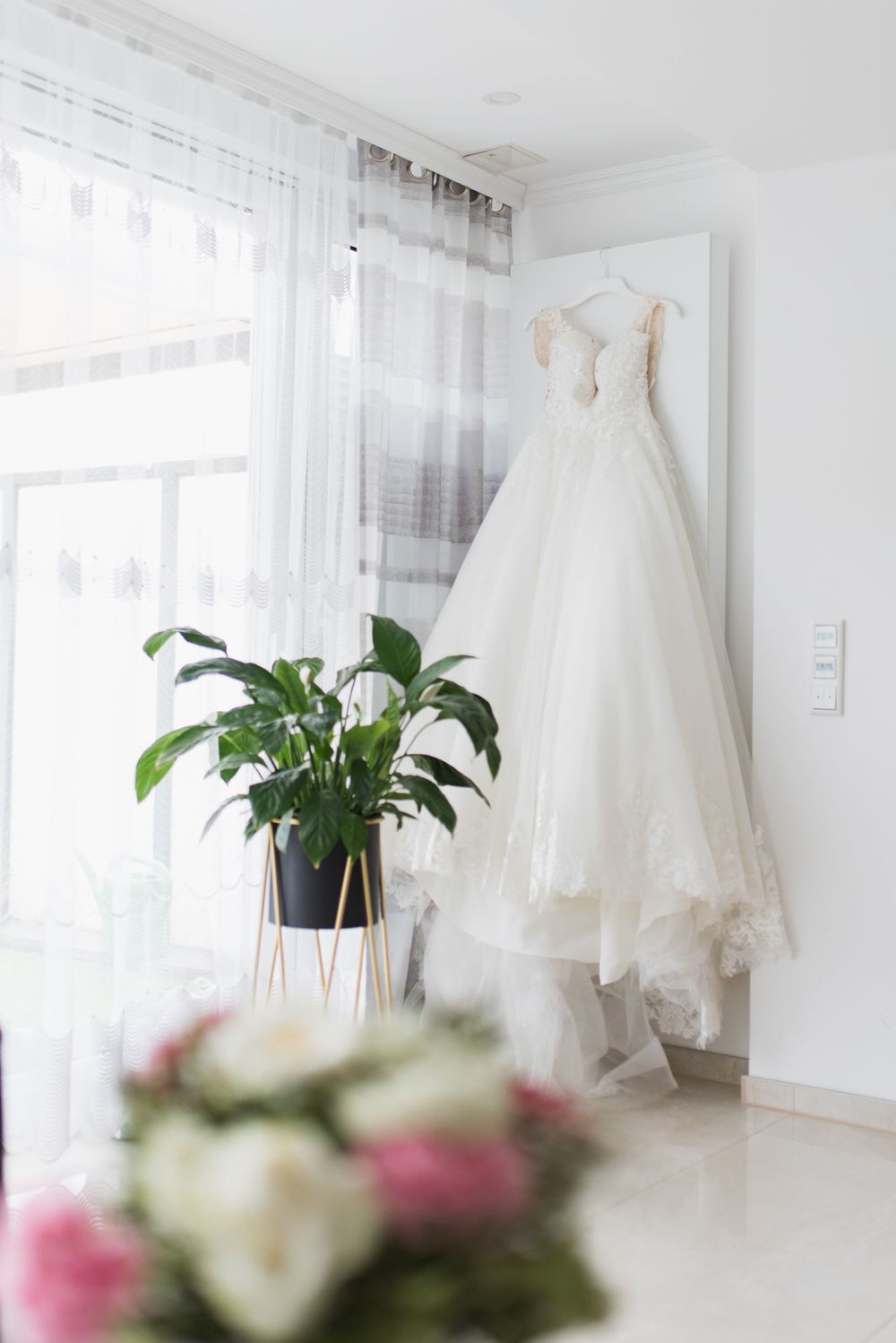 Elegant white wedding dress hanging against sheer curtains with potted plant and floral decor.