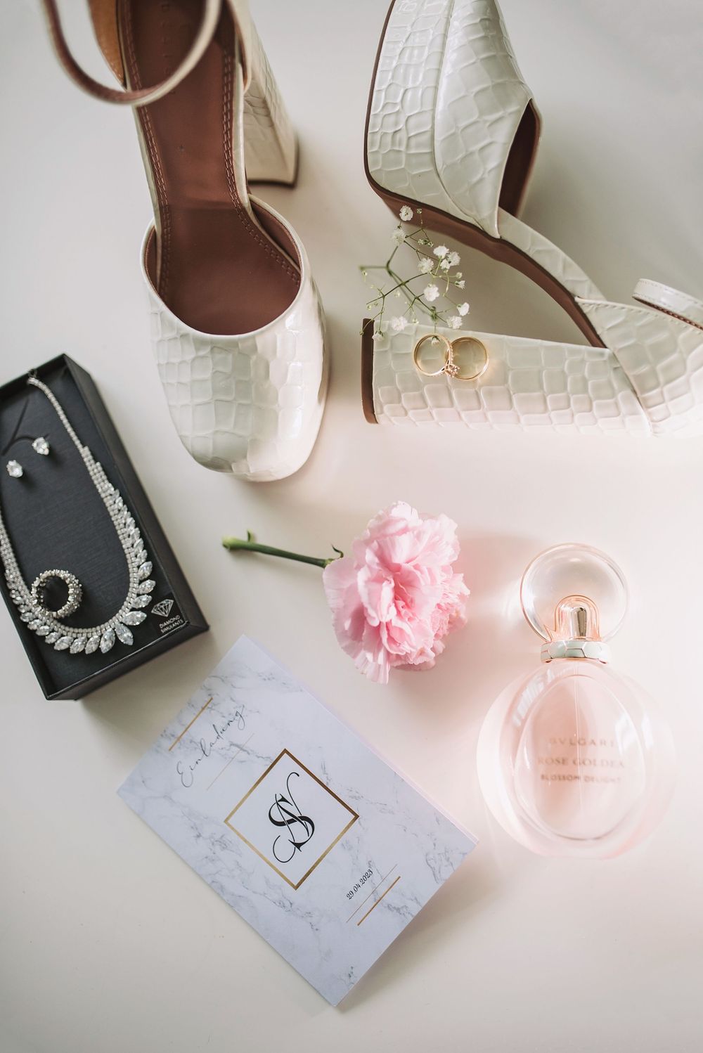 Elegant bridal accessories including white shoes, pink flower, perfume bottle and jewelry displayed on light surface.
