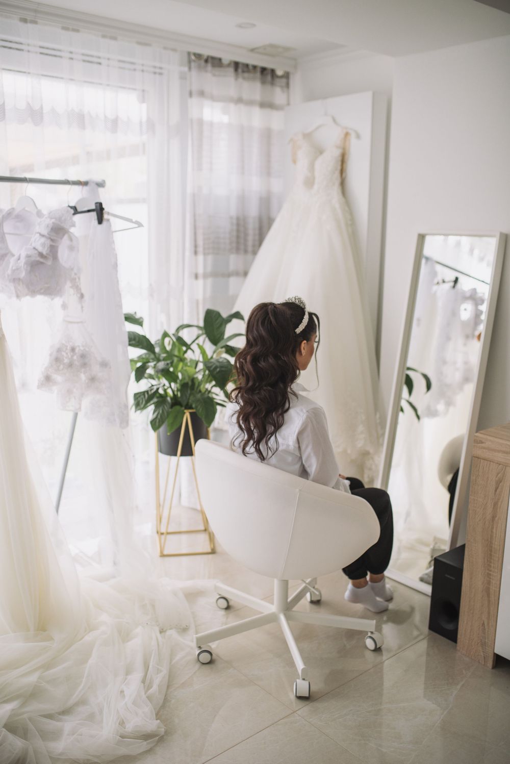 Wedding dress hanging in bright room with office chair and potted plant near window.