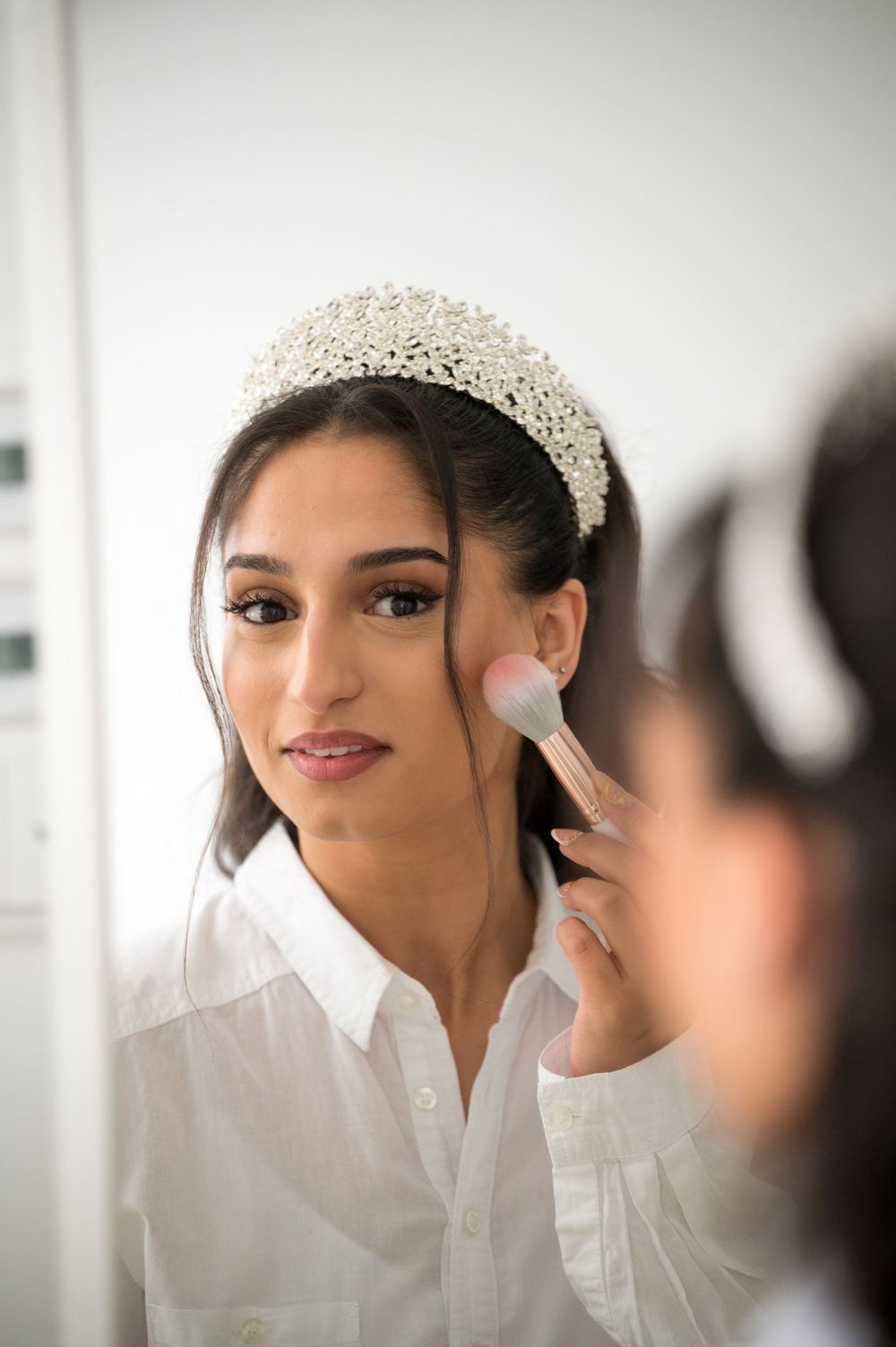 Elegant bridal preparation with crystal headpiece and makeup application in natural light.