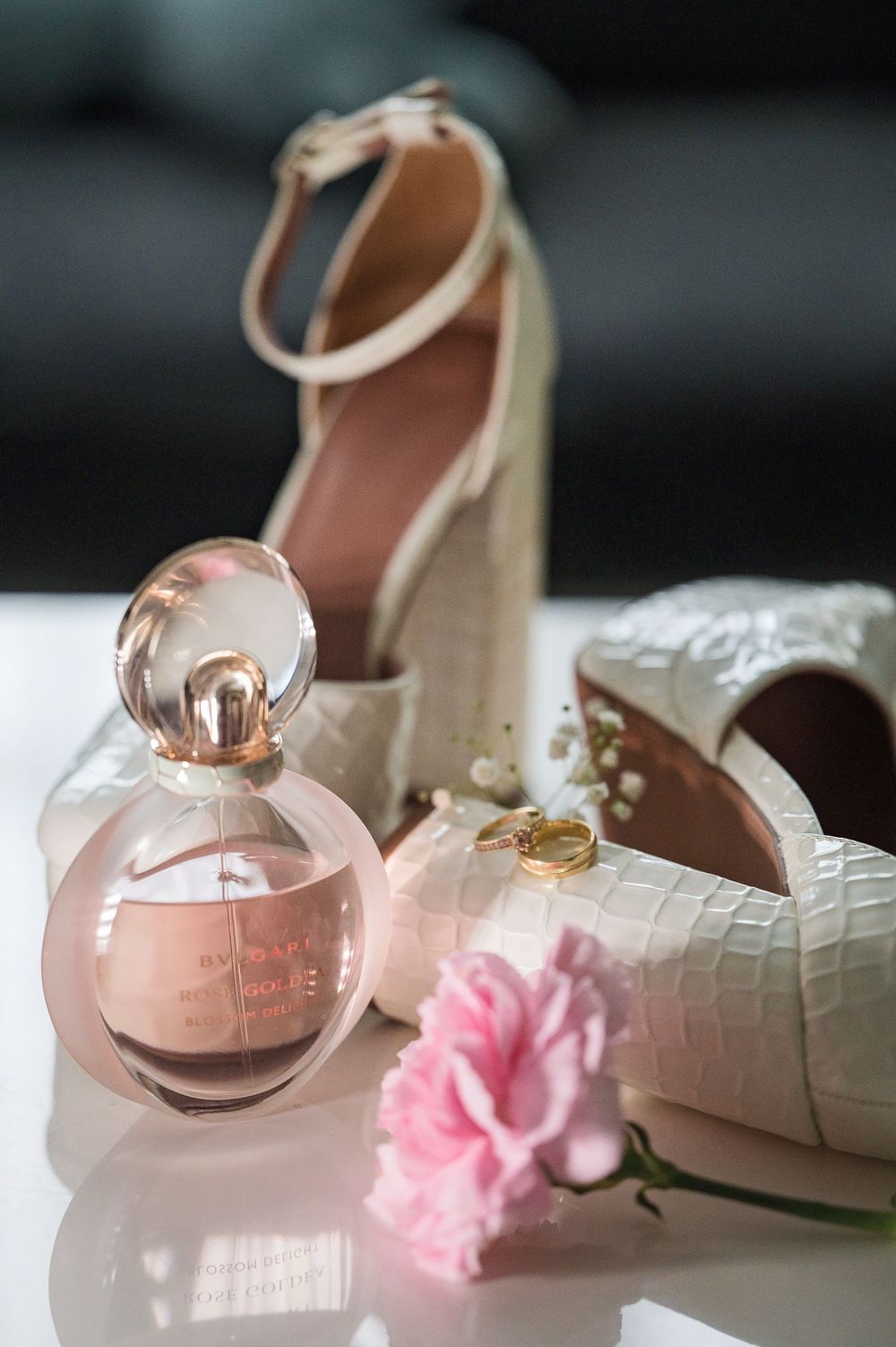 White bridal shoes with croc texture next to pink perfume bottle and delicate pink flower.
