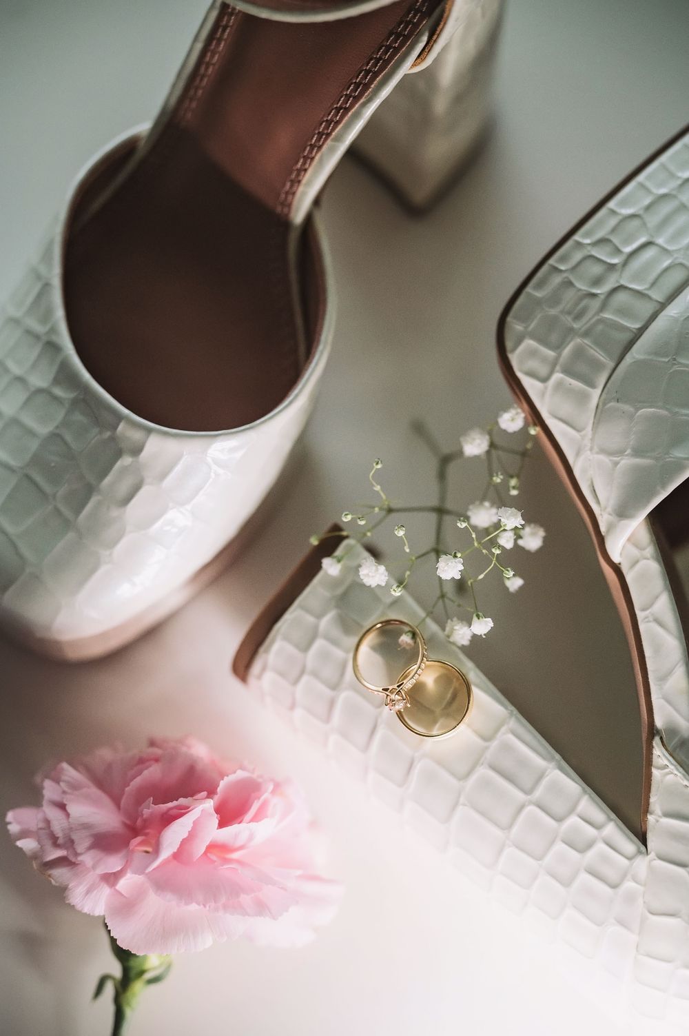 White textured bridal shoes with wedding rings and pink flower detail.