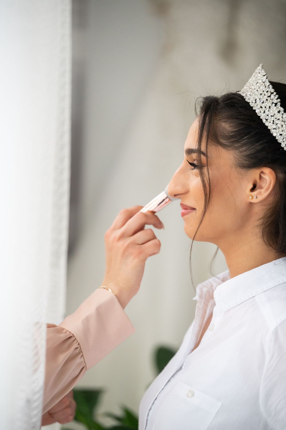 Wedding makeup application in side profile with crystal crown and white attire.