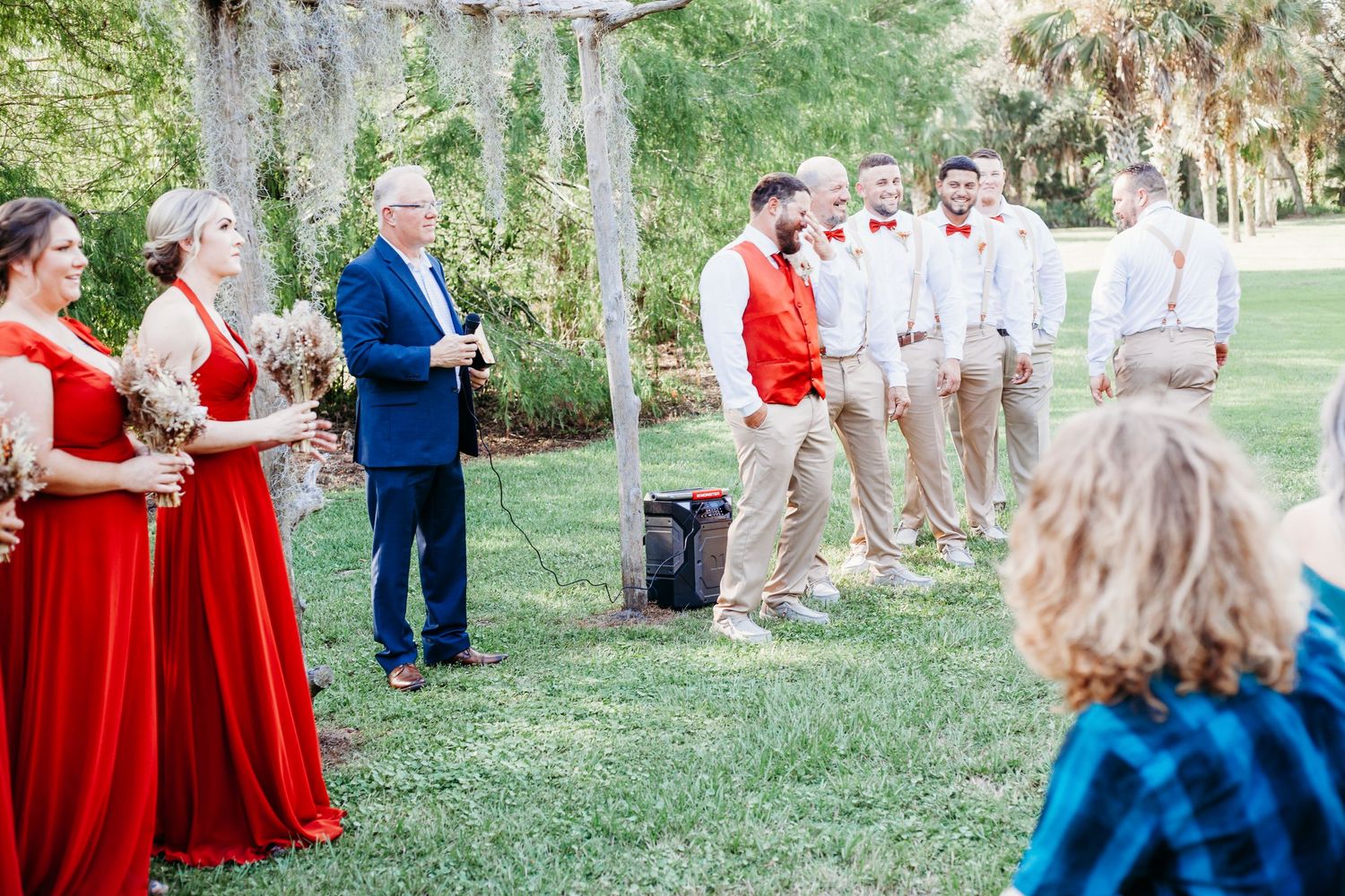 A wedding party stands on a lawn with bridesmaids in red dresses and groomsmen in khaki under Spanish moss.
