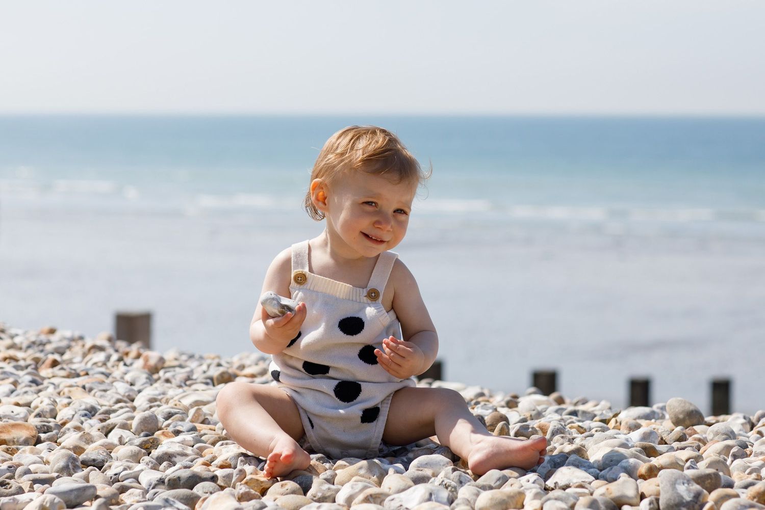 Small child in polka dot outfit sitting happily on a pebble beach by the ocean.