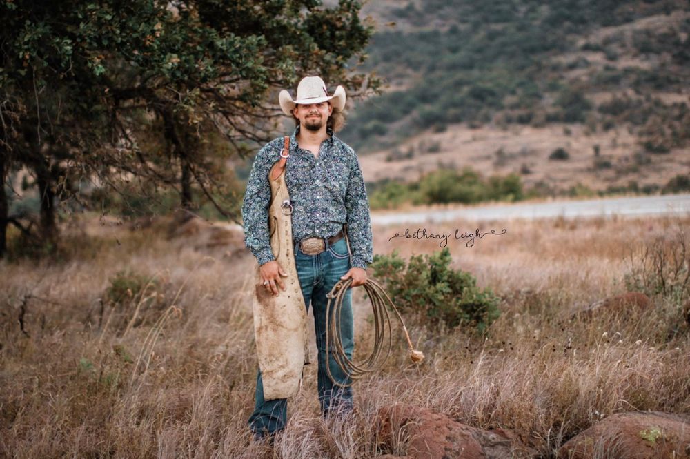 Cowboy with rope and traditional western wear standing in rural landscape.