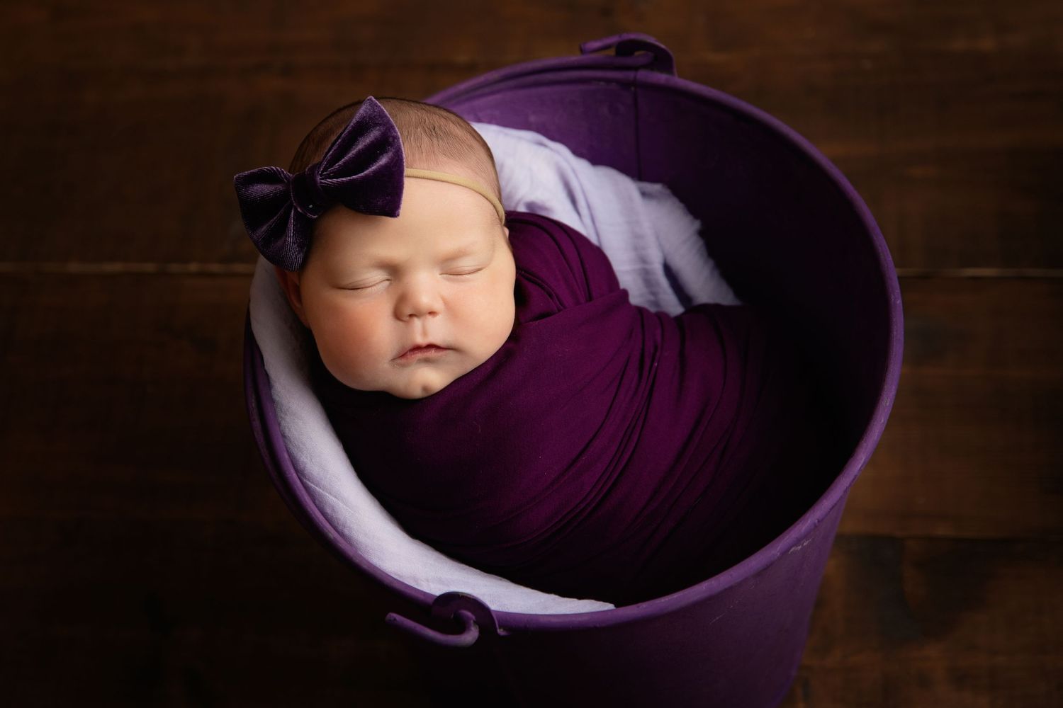 Sleeping newborn wrapped in purple fabric inside purple bucket with matching bow.