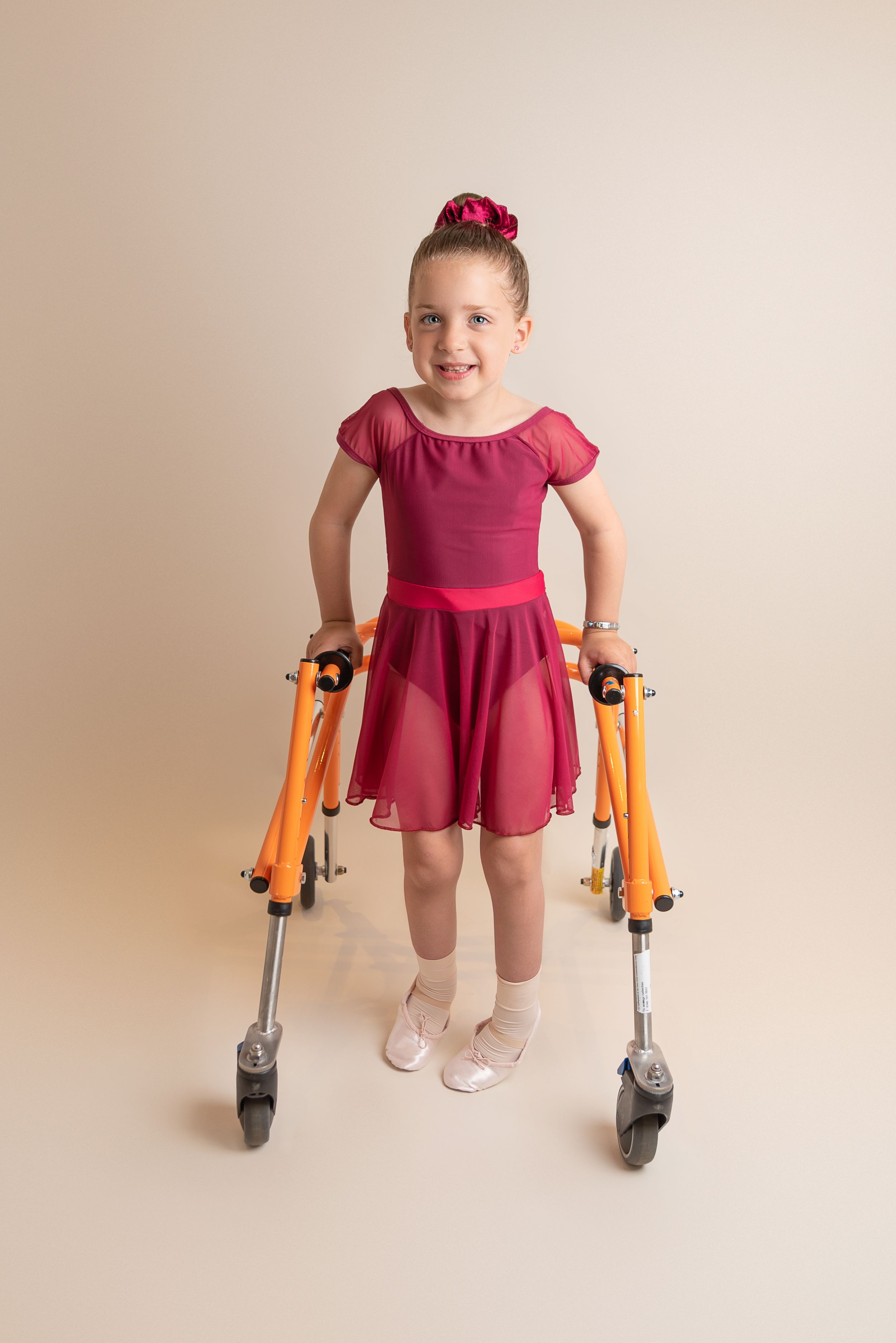 Young dancer with cerebral palsy in a red dress stands with orange mobility walker against beige background.