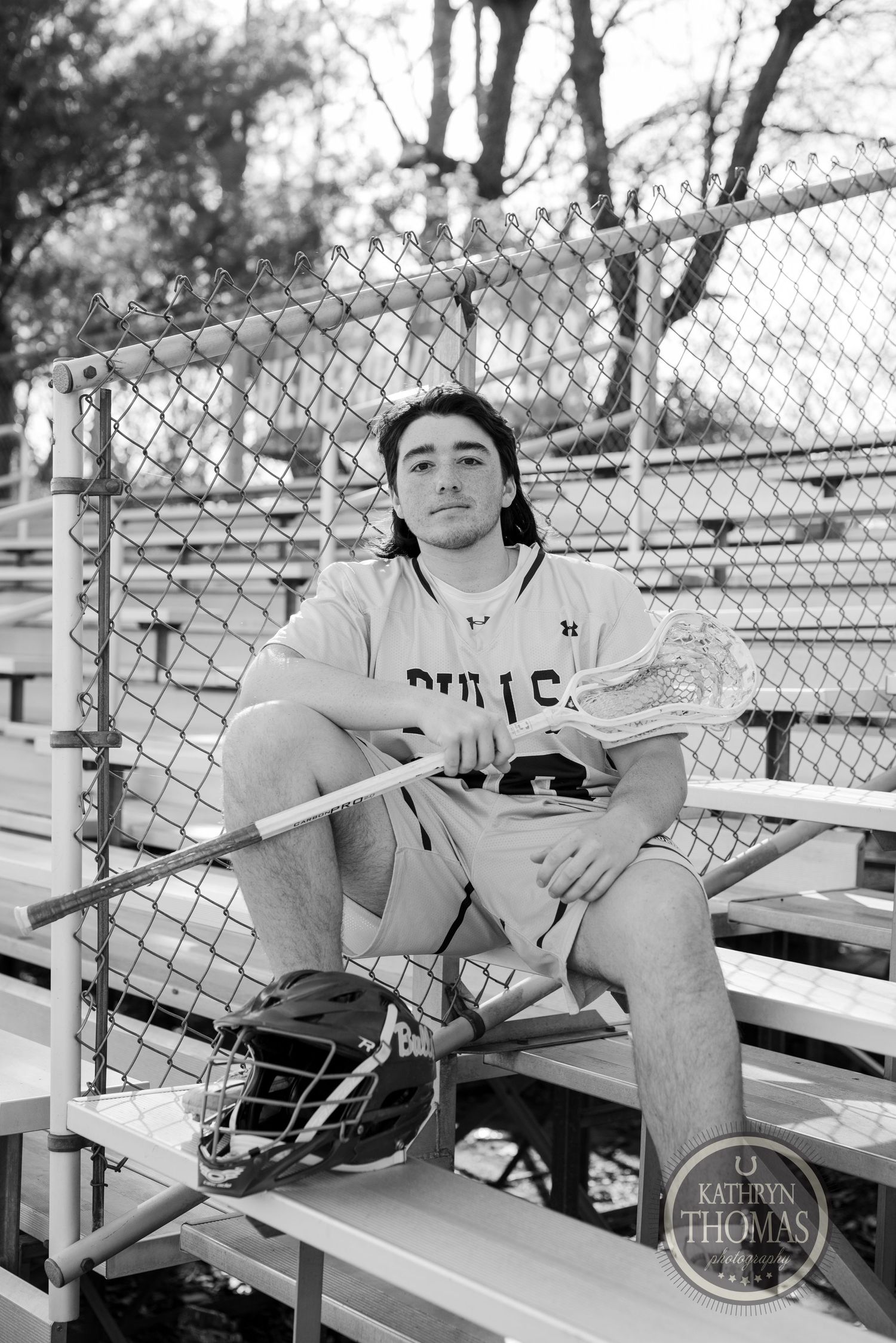 Athletic player in white uniform sits on bleachers with baseball equipment in black and white photo.