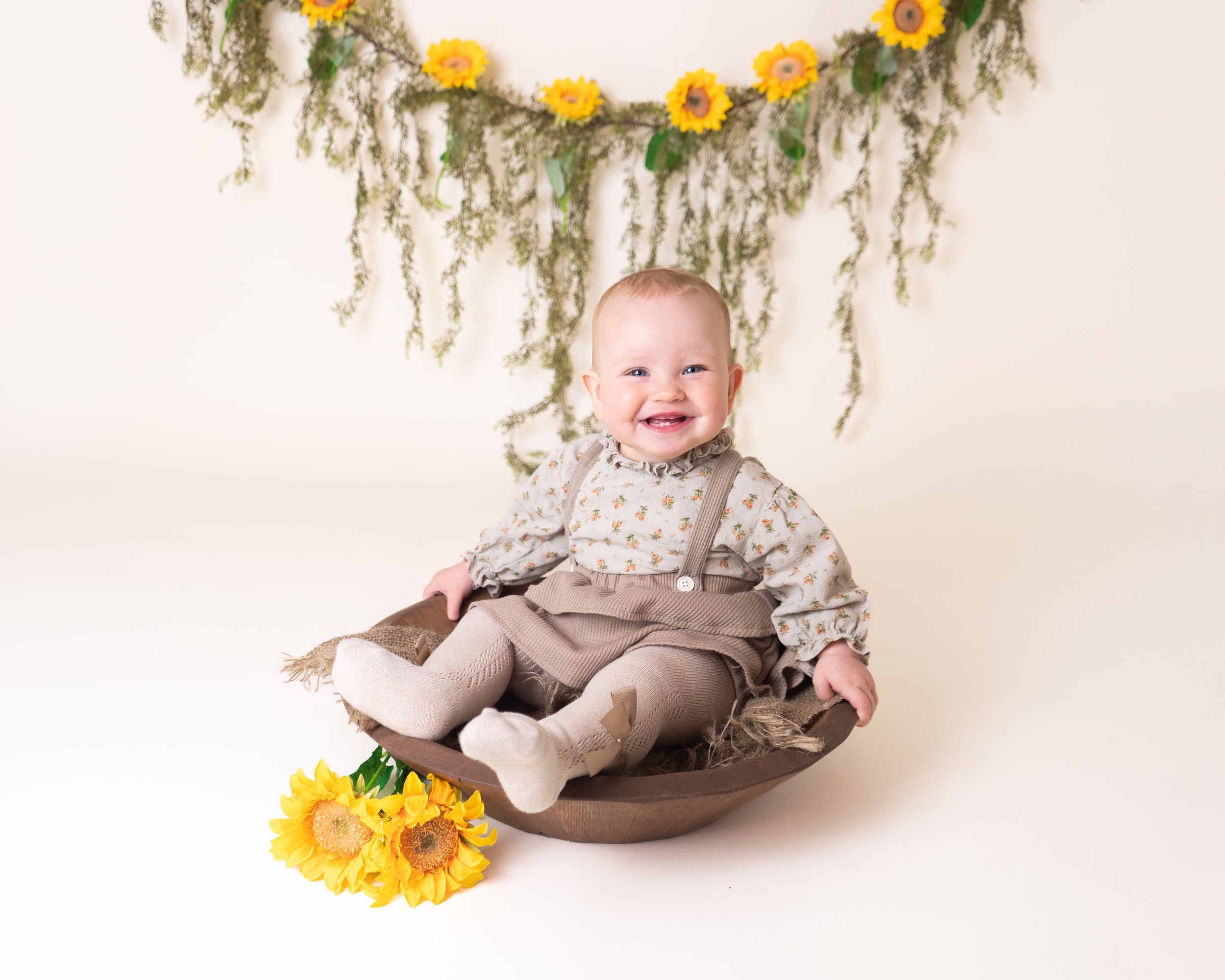 first birthday baby girl sat smiling in a wooden bowl with sunflowers, by newborn photographer in doncaster.