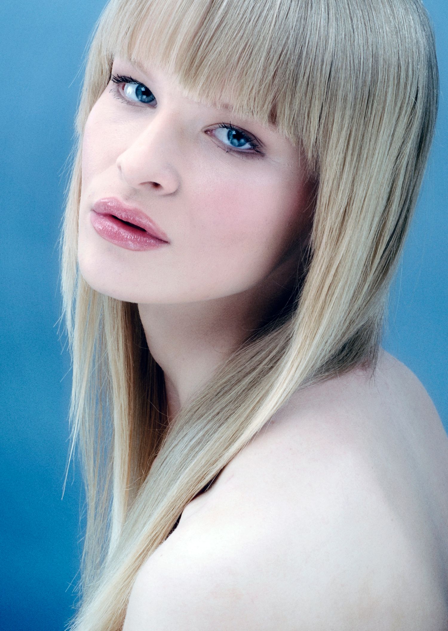 Beauty portrait with long blonde hair and bangs against blue background.