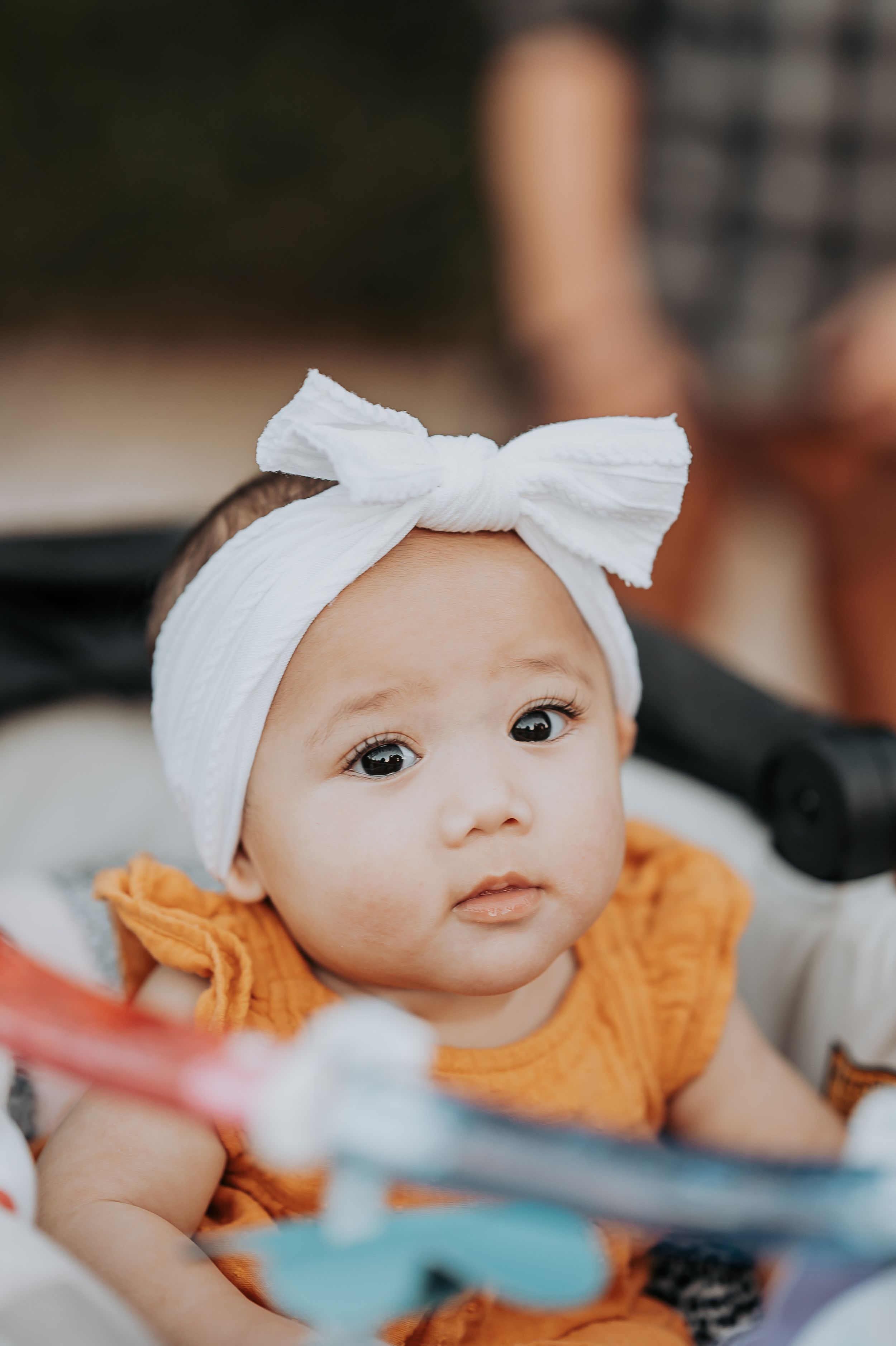 Baby wearing white bow headband and orange dress looking at camera with curious expression.