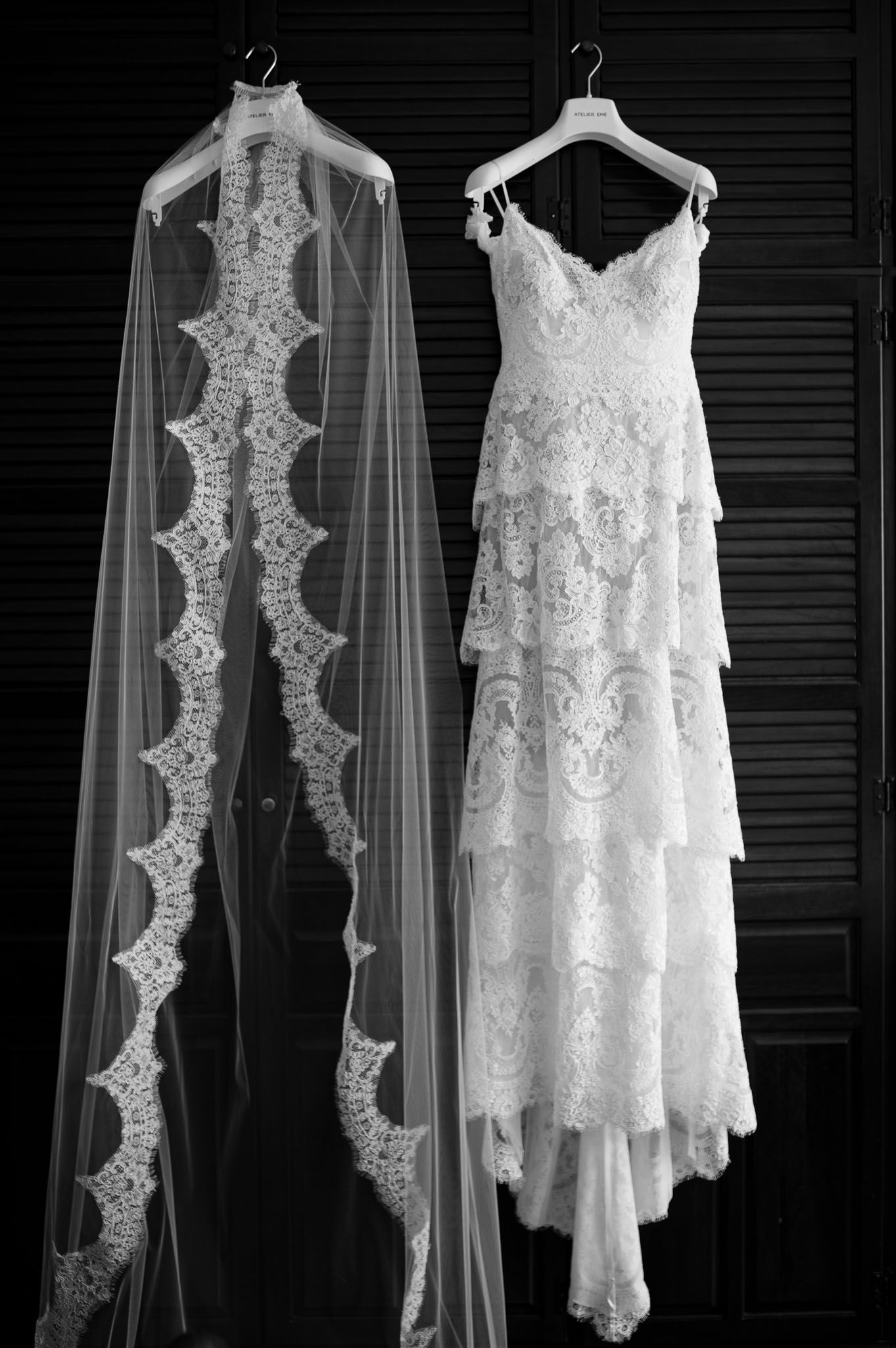 Black and white photo of a wedding dress and veil hanging side by side showing intricate lace details.