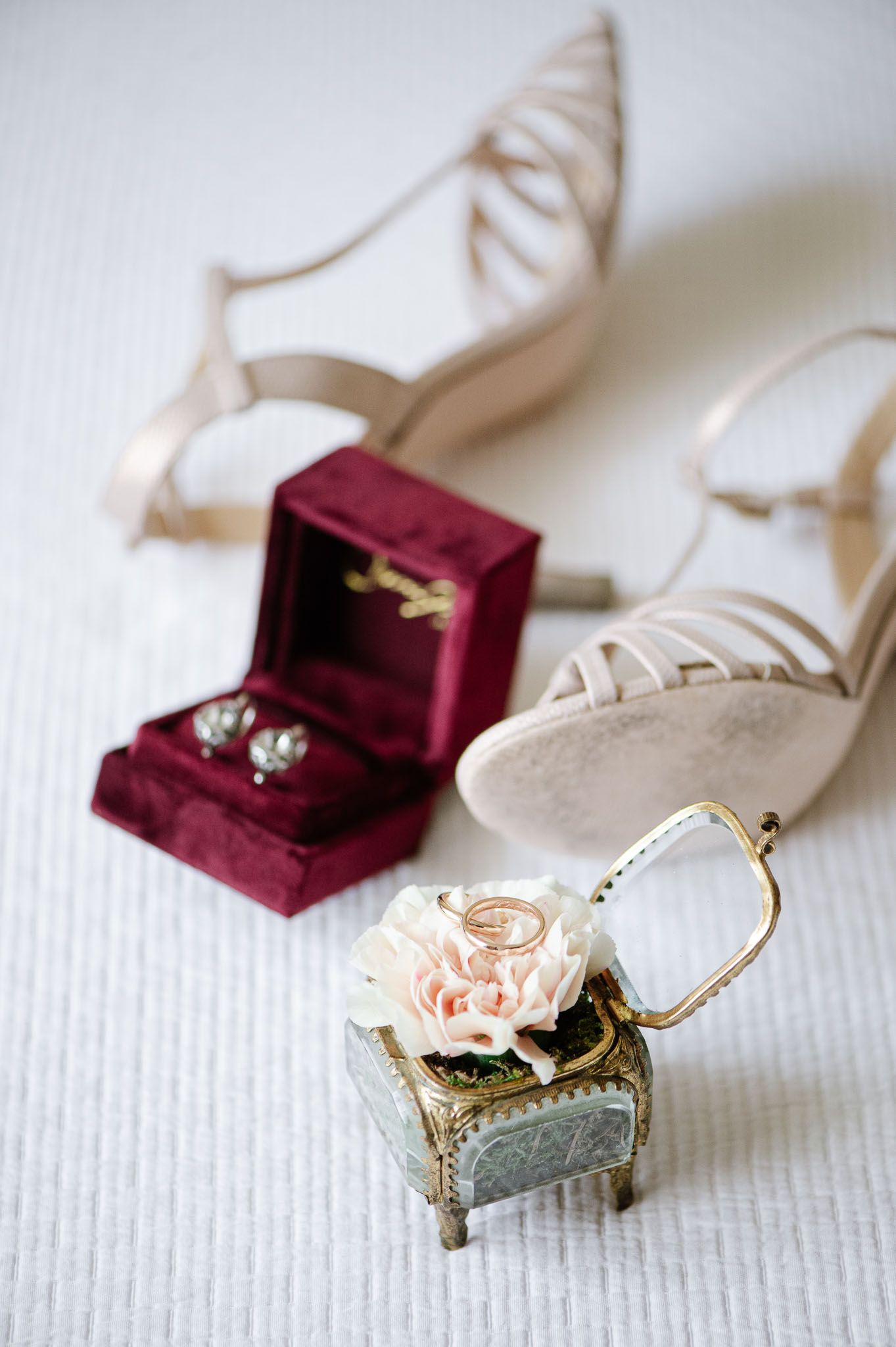 Wedding accessories including ivory shoes, burgundy ring box, and delicate floral jewelry display box on white surface.