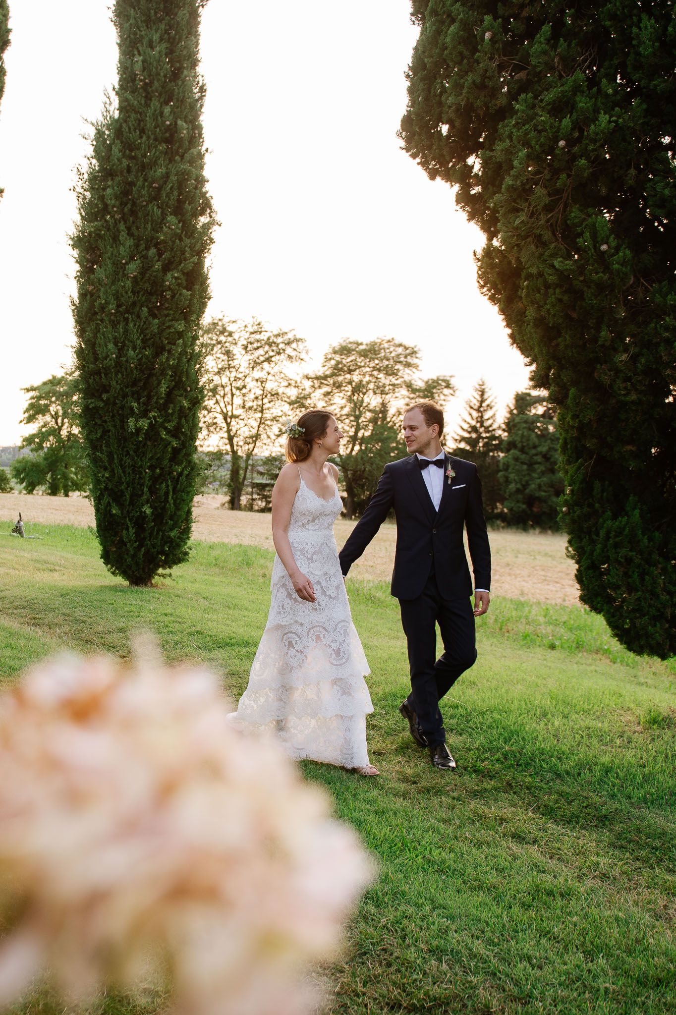 Couple in wedding attire walking between tall cypress trees at sunset.