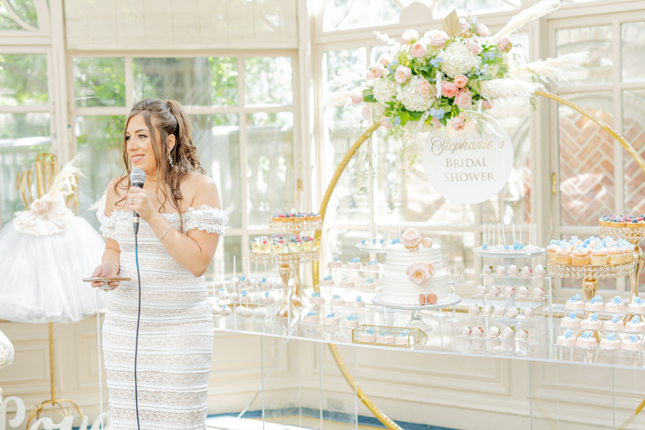 Bride in white dress speaking into microphone in elegant greenhouse venue.