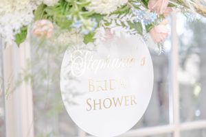 White and gold bridal shower sign hanging from floral arrangement with soft lighting.