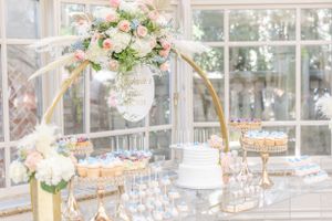 Luxurious dessert table with gold arch and floral arrangements in a sunlit conservatory.