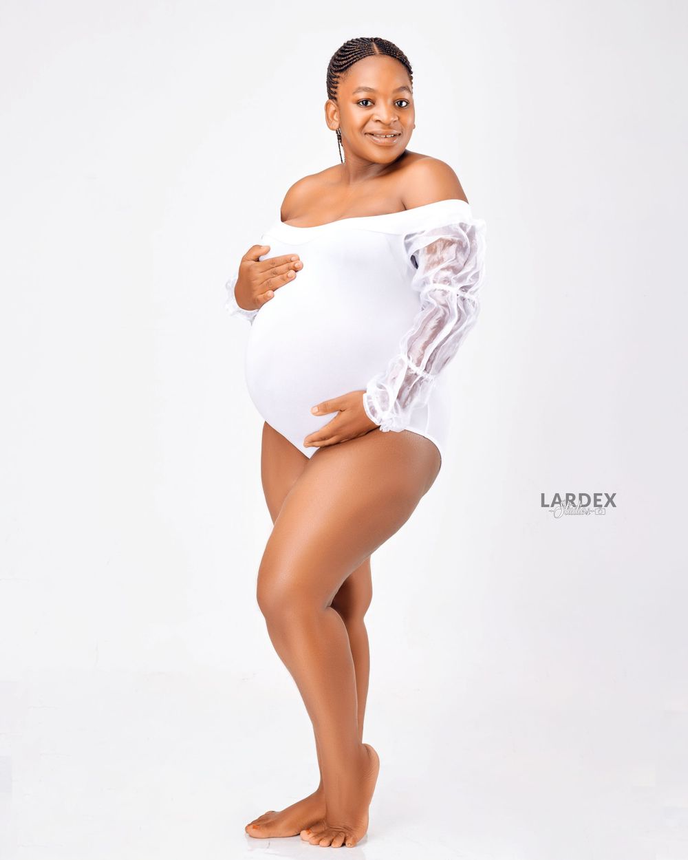 Maternity photo of someone in a white lace bodysuit posing against light background.