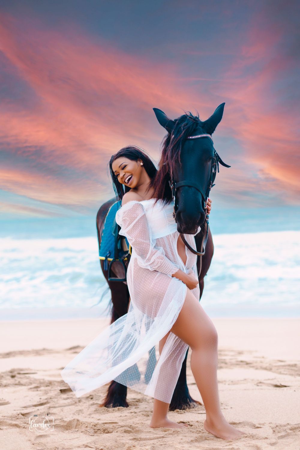Woman in flowing white dress poses with dark horse on beach during sunset.