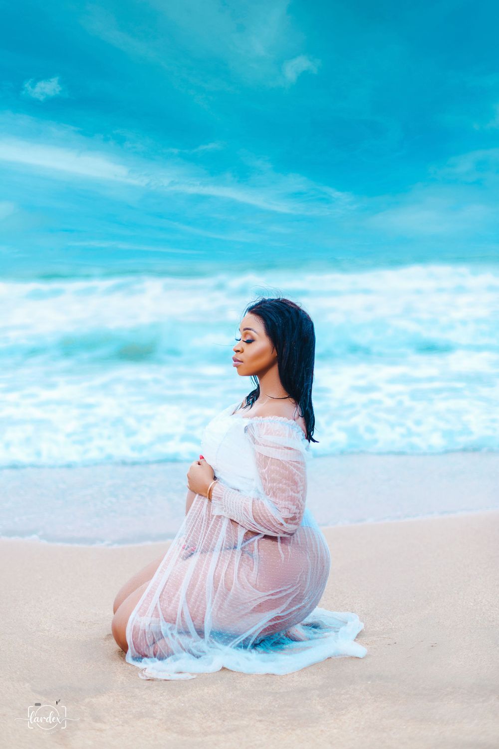 Woman in white lace dress kneels peacefully on beach under blue sky.