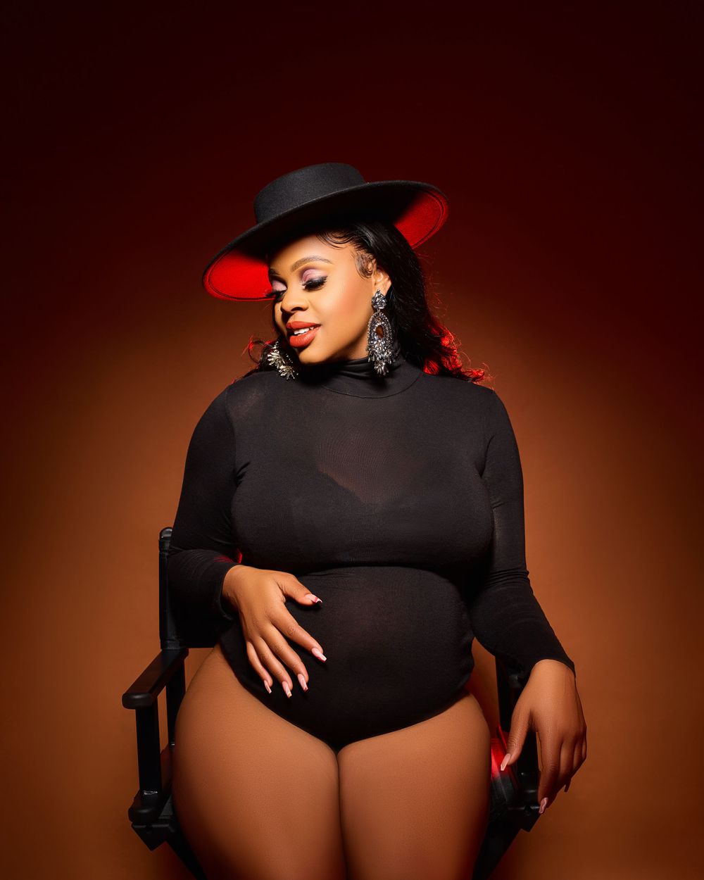 Artistic maternity photo featuring a dramatic black bodysuit and hat against a deep red backdrop.
