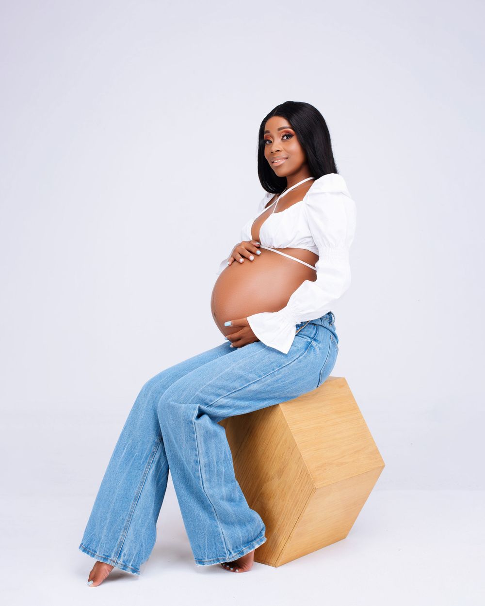 Modern maternity photo with casual white top and jeans while sitting on wooden cube.