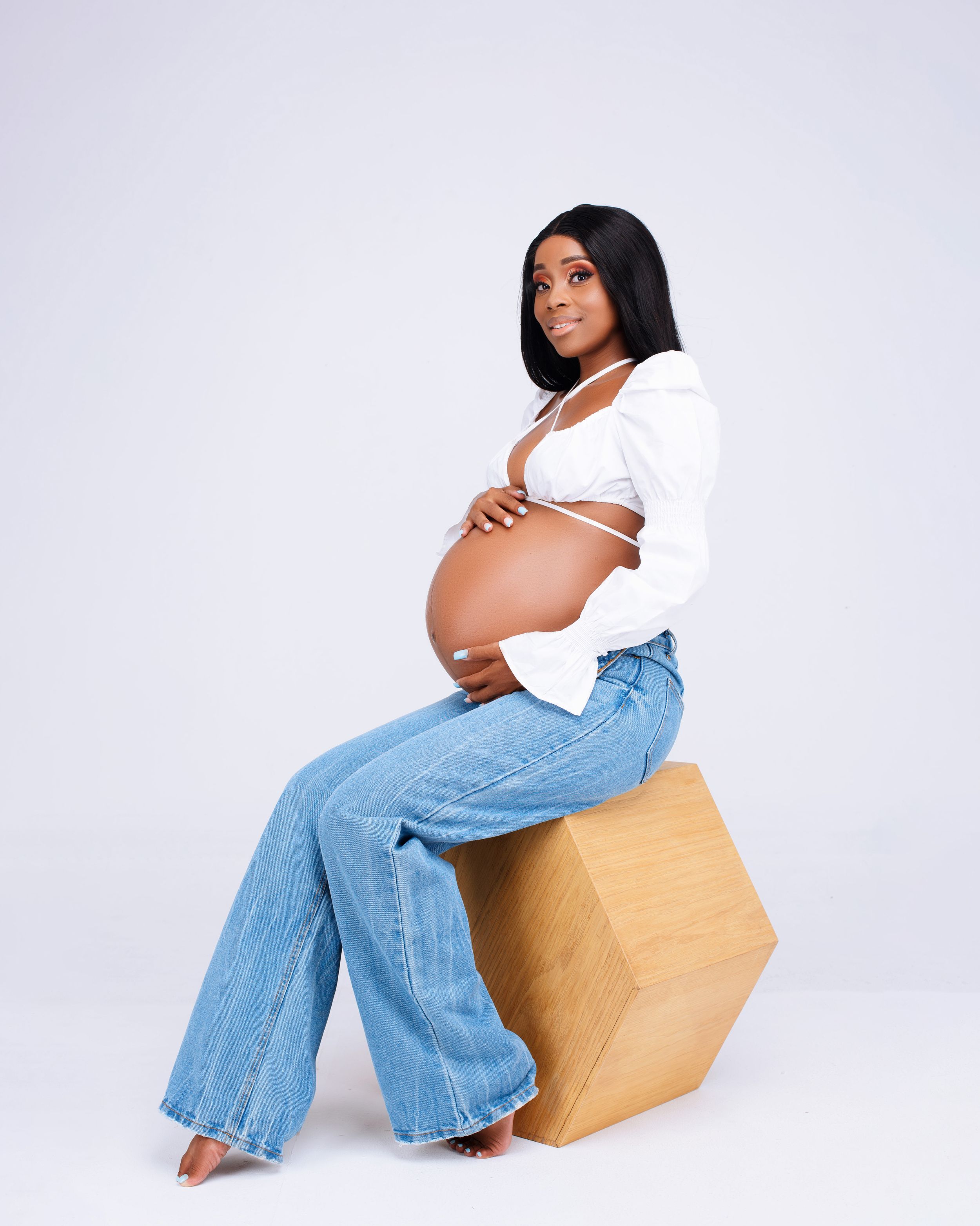 Modern maternity photo with casual white top and jeans while sitting on wooden cube.