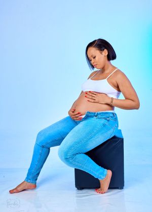 Expectant mother in casual denim and white tank top poses on black cube against blue gradient backdrop.