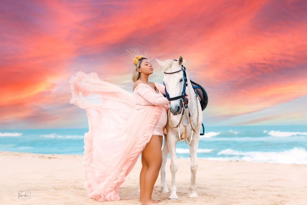 Woman in pink flowing dress poses with white horse on beach at sunset.