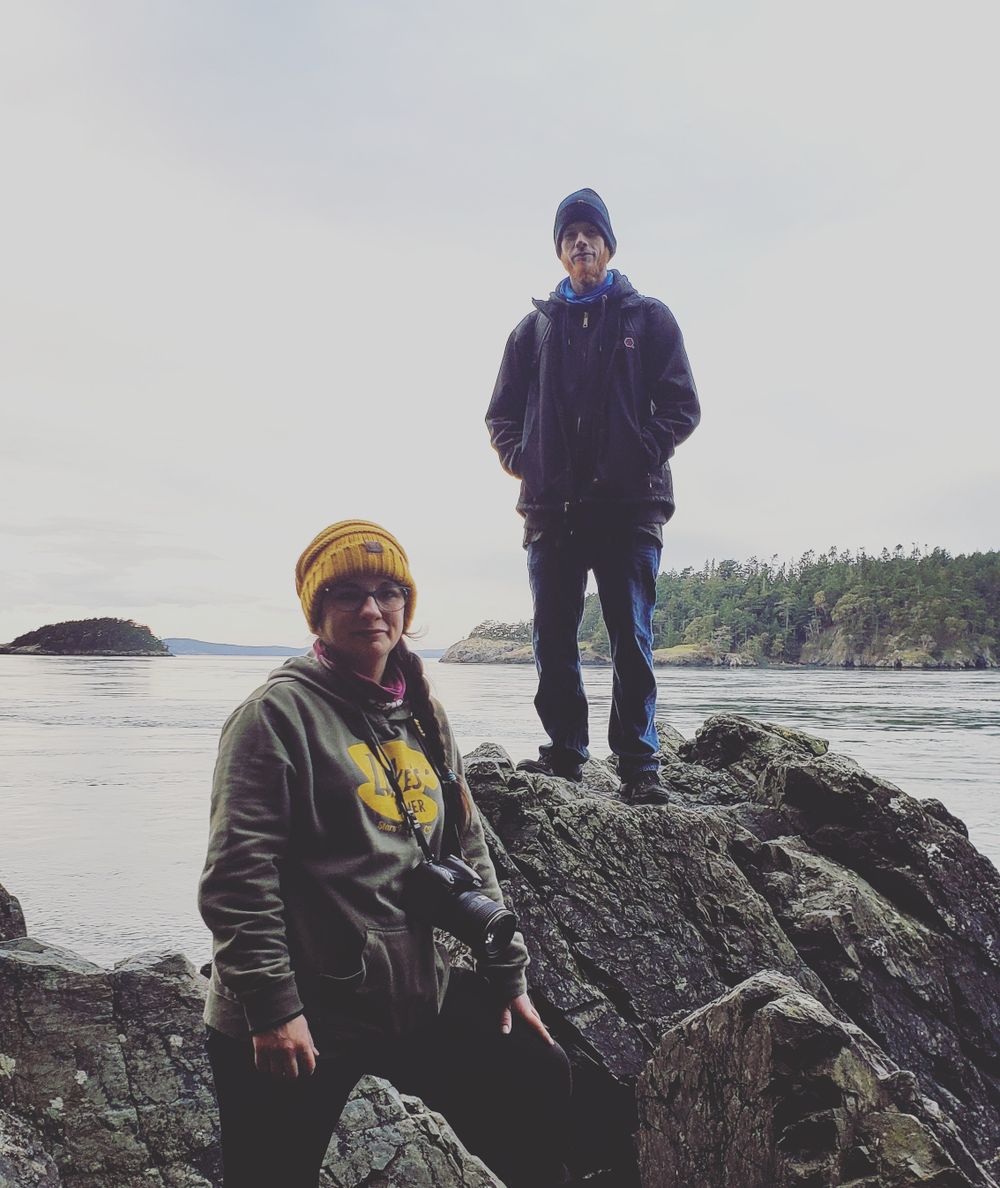 Two people bundled in winter gear on coastal rocks overlooking calm water and islands