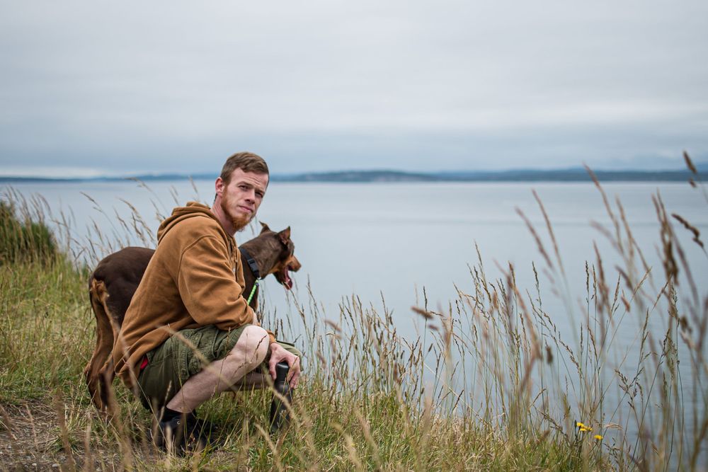 Man and his brown dog squatting on a grassy cliff overlooking the ocean. Whidbey island scenic portrait