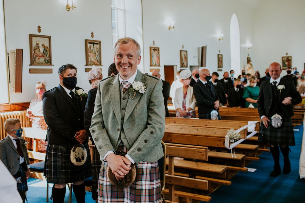Wedding attendee in traditional Scottish kilt and green jacket standing in church aisle.