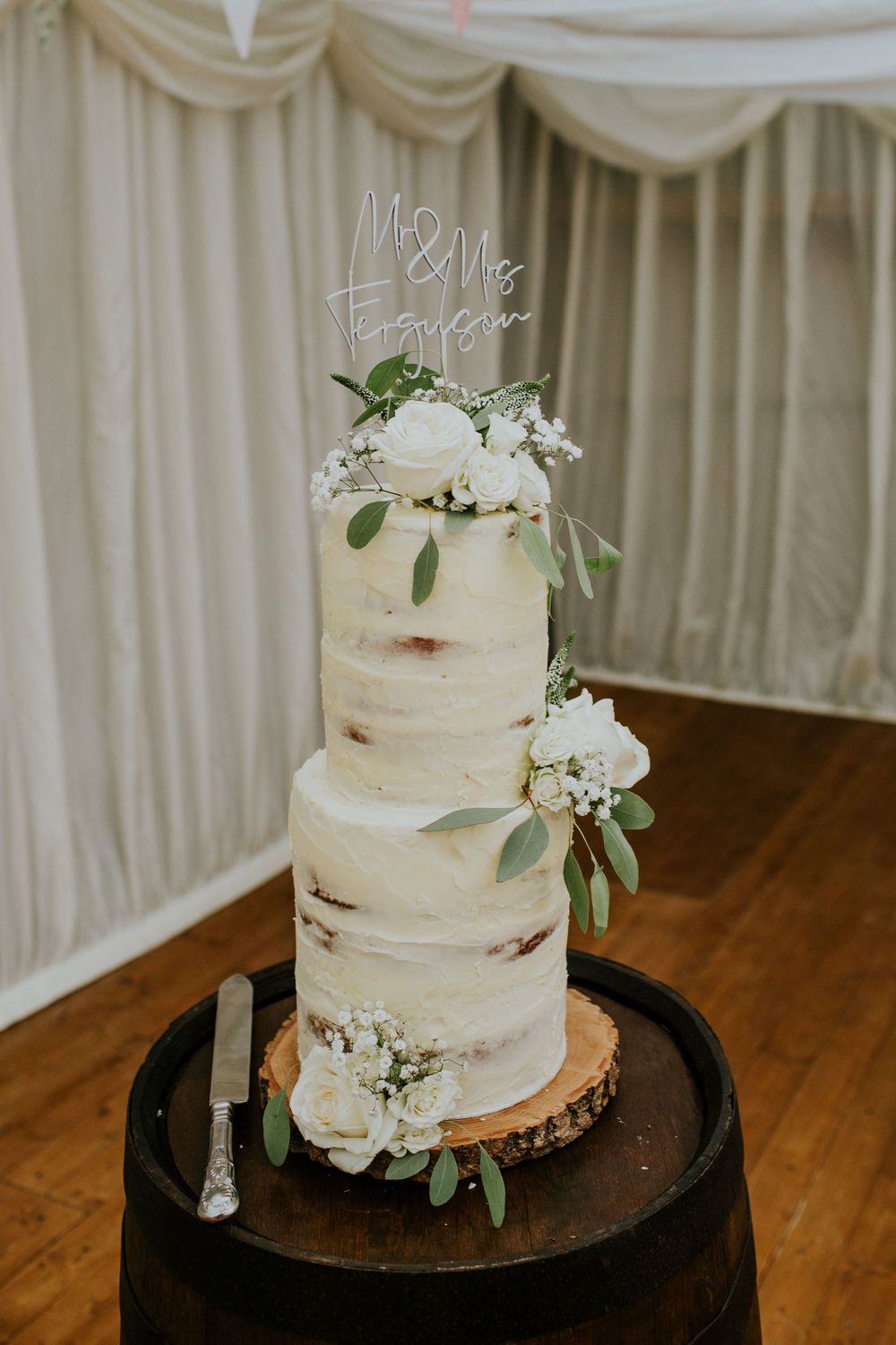 Semi-naked white wedding cake decorated with fresh flowers and greenery on a wooden barrel stand.