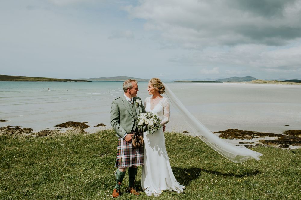 A Scottish wedding couple in kilt and white dress pose by the scenic coastline.