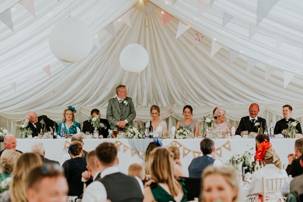 Wedding reception guests seated at a long head table under an elegant white draped tent ceiling.