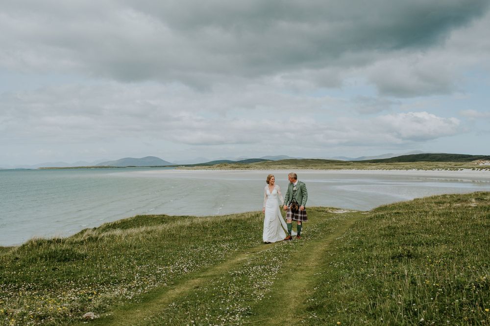A couple stands on a grassy path overlooking a pristine beach and mountains.