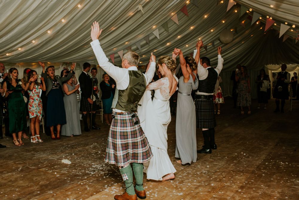 Energetic wedding dance featuring traditional Scottish kilts in a decorated marquee tent.