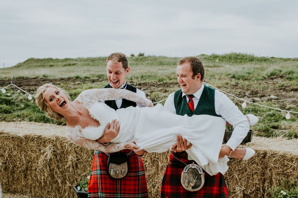 Groomsmen in kilts playfully carry bride in outdoor countryside setting.
