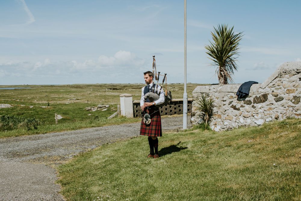 Bagpiper in traditional Scottish dress performs on grassy coastal path.