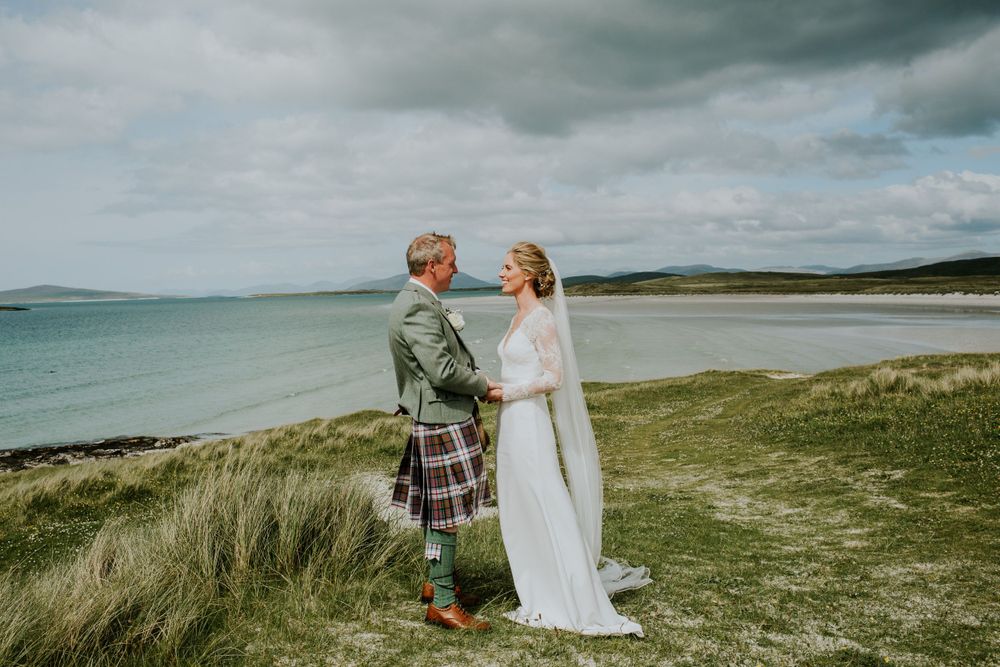 A couple in wedding attire stands together on windswept coastal grassland.