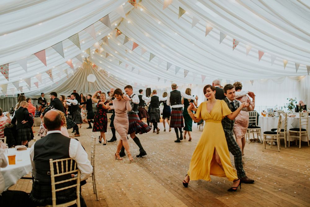 Wedding guests dancing in an elegant white marquee decorated with pastel bunting and lights.