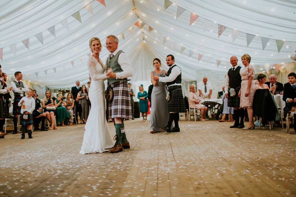 Wedding couple dancing in Scottish attire under white tent with fairy lights and bunting.