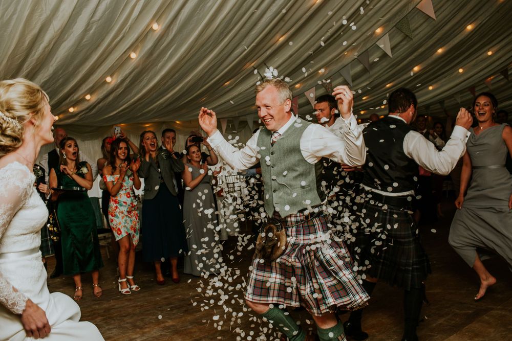 A joyous celebration at a Scottish wedding with kilts and confetti in a draped tent reception venue.