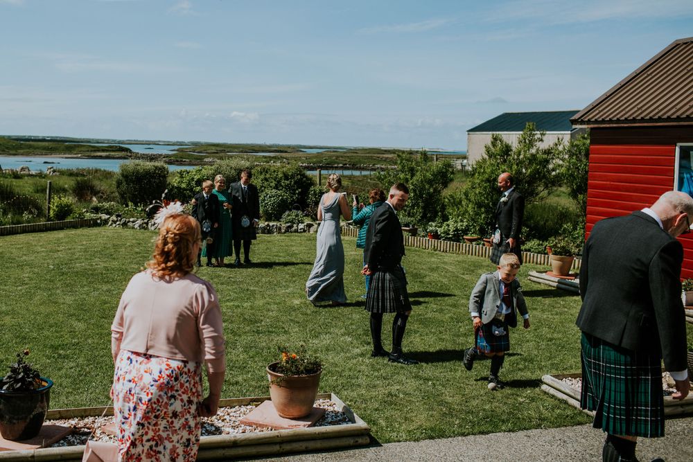 Wedding guests gather on a grassy lawn with coastal views and red building in background.