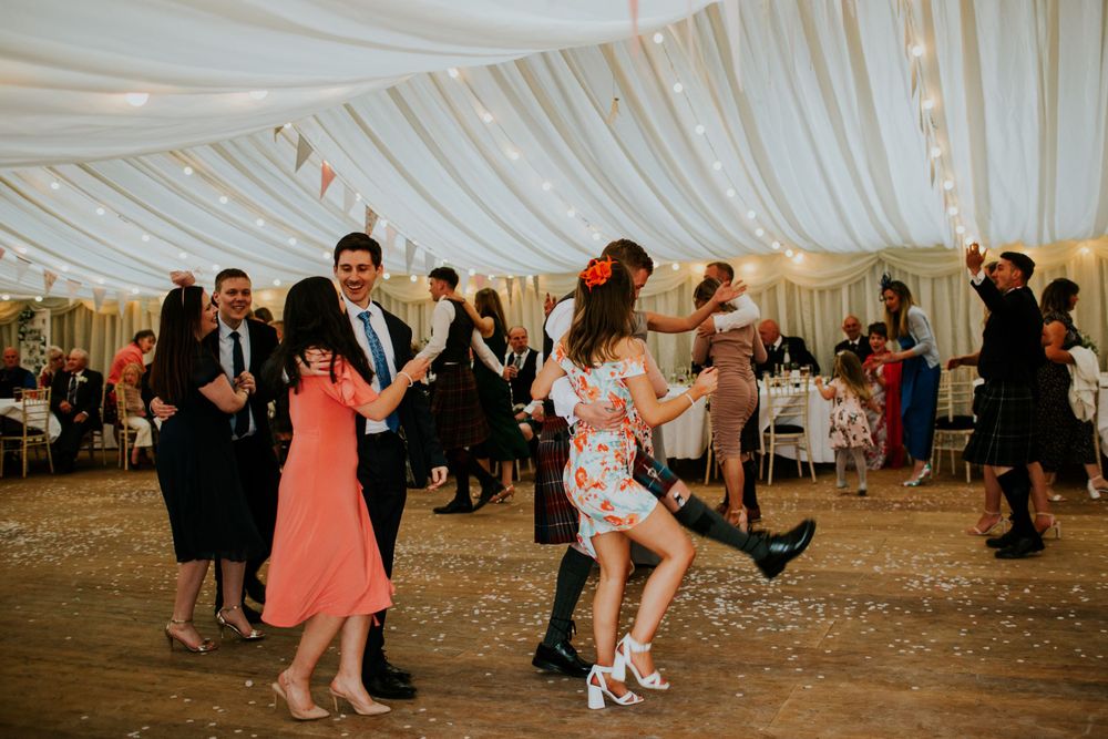 Lively dancing at a wedding reception under white draped ceiling with festive lighting.