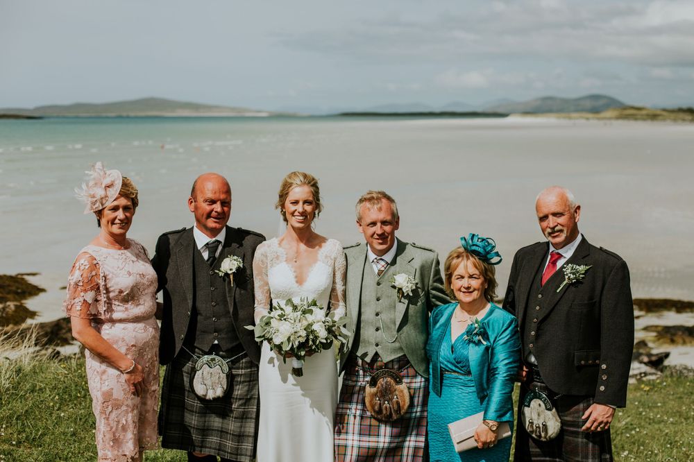A wedding party in traditional Scottish attire poses on a grassy beach overlook.